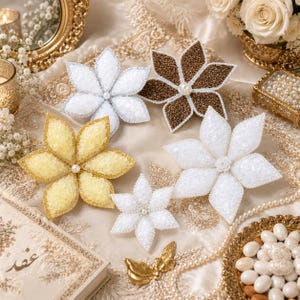 May include: A collection of five decorative star-shaped ornaments featuring textured surfaces in white, gold, and brown. Each ornament has six pointed petals, a central pearl-like bead, and glittery edges. The set includes varying sizes of these floral-inspired pieces.