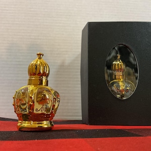 May include: A gold-colored perfume bottle with a crown-shaped top and ornate detailing. The bottle is displayed next to a black box with an oval window showcasing the bottle. The background features a red and black plaid pattern.