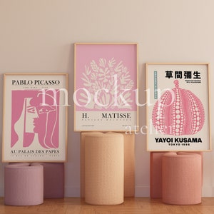Könnte beinhalten: Drei gerahmte Kunstdrucke mit rosa Hintergründen und weißen Designs. Die Drucke befinden sich auf einem Regal mit zwei rosa und einem beigefarbenen zylindrischen Ständern. Die Drucke zeigen die Namen Pablo Picasso, H. Matisse und Yayoi Kusama. Der Druck von Yayoi Kusama zeigt den japanischen Text "草間彌生" und die Wörter "Yayoi Kusama" und "Tokyo 1998".