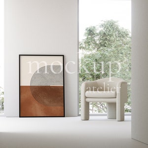 May include: A white armchair with a brown and white abstract geometric print in a black frame hanging on a white wall. The chair is in front of a window with a view of green trees.