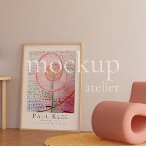 May include: A framed print of a colorful abstract painting by Paul Klee titled "Blumenwy - 1914" hangs on a light beige wall. A pink armchair sits in front of the print, and a green lamp sits on a wooden table.