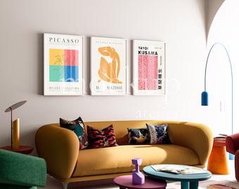 Op de afbeelding: Drie ingelijste kunstprints met minimalistische ontwerpen in een woonkamer. De prints tonen de namen "Picasso", "H. Matisse" en "Yayoi Kusama" in zwarte tekst. De prints hangen aan een witte muur boven een gele bank met kleurrijke kussens.