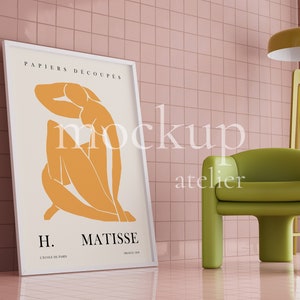 May include: A framed poster with a white background and a yellow abstract figure. The poster is titled "PAPIERS DECOUPES" and "H. MATISSE". The poster is leaning against a pink tiled wall next to a green armchair and a yellow floor lamp.