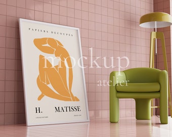 Op de afbeelding: Een ingelijste poster met een witte achtergrond en een gele abstracte figuur. De poster is getiteld "PAPIERS DECOUPES" en "H. MATISSE". De poster leunt tegen een roze betegelde muur naast een groene fauteuil en een gele staande lamp.
