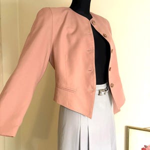 Puede incluir: Una blazer rosa claro con cuello redondo y tres botones. La blazer se combina con una falda gris y una blusa negra. El conjunto se exhibe en un maniquí.
