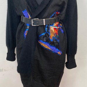 May include: A black knit cardigan with a long, open front and a wide collar. The cardigan has a sequined design on the front and a black leather belt.