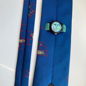 Puede incluir: Una corbata de seda azul con un patrón repetido de coches rojos sobre un fondo blanco. La corbata tiene un reloj negro con una esfera colorida unido a ella. La corbata tiene una etiqueta negra que dice "Made in Germany".