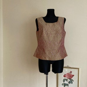 May include: A sleeveless, gold-colored top with a pink floral pattern and sequins. The top is on a black mannequin.