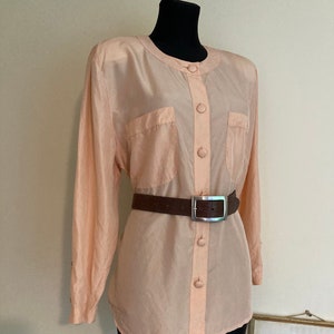 May include: A peach-colored silk blouse with long sleeves and a button-up front. The blouse has two patch pockets and a brown leather belt with a silver buckle.