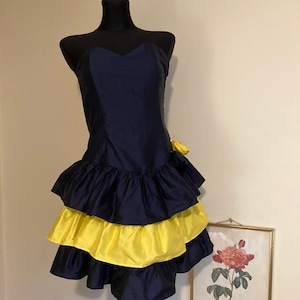 Pode incluir: Um vestido curto sem alças, preto e amarelo, com uma saia de babados. O vestido tem um decote em coração e um laço amarelo no lado.