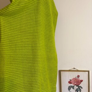 May include: A bright lime green fabric with a textured, dotted pattern. The fabric is draped, with a glimpse of a framed botanical print in the background. The print features a pink rose illustration.