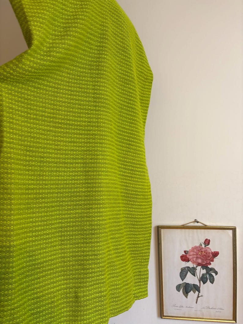 May include: A bright lime green fabric with a textured, dotted pattern. The fabric is draped, with a glimpse of a framed botanical print in the background. The print features a pink rose illustration.