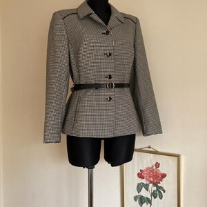 Może przedstawiać: Czarno-biała marynarka w kratę typu „pieski” z czarnym paskiem i złotą klamrą. Marynarka ma kołnierz i zapięcie na guziki.