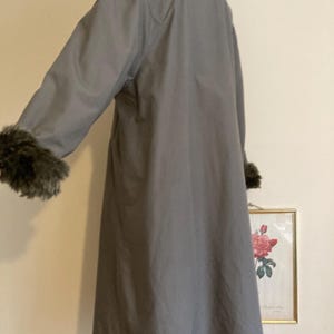 May include: A long gray coat with a faux fur collar and cuffs. The coat has a simple design and is made of a smooth fabric.