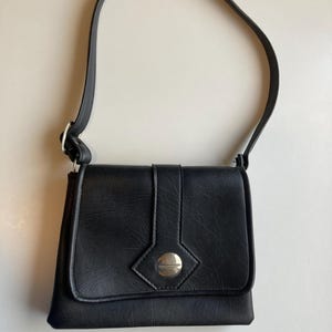 Pode incluir: Bolsa de ombro preta com fecho de aba e bot&atilde;o prateado. A bolsa tem uma al&ccedil;a longa e formato retangular. A bolsa &eacute; feita de um material texturizado.