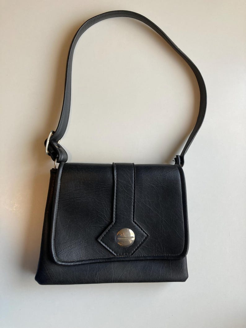 Pode incluir: Bolsa de ombro preta com fecho de aba e bot&atilde;o prateado. A bolsa tem uma al&ccedil;a longa e formato retangular. A bolsa &eacute; feita de um material texturizado.