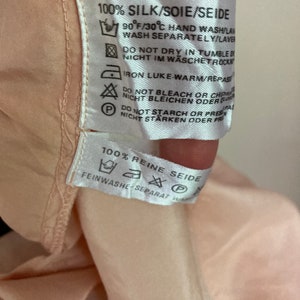 May include: A white care label with black text and symbols. The label reads "100% Silk/Soie/Seide" and includes instructions for hand washing, drying, ironing, and bleaching. The label also reads "100% Reine Seide" and includes instructions for delicate washing.