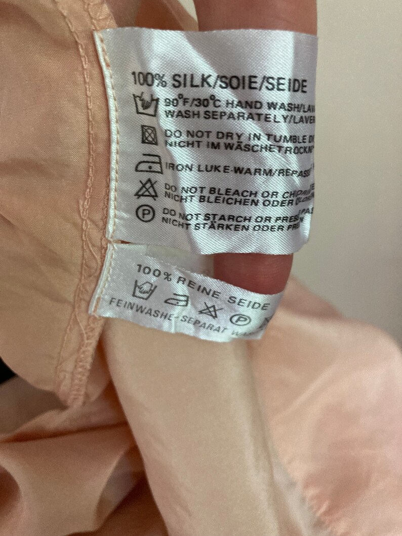 May include: A white care label with black text and symbols. The label reads "100% Silk/Soie/Seide" and includes instructions for hand washing, drying, ironing, and bleaching. The label also reads "100% Reine Seide" and includes instructions for delicate washing.