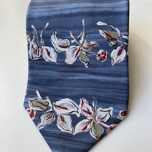 Op de afbeelding: Een blauwe zijden stropdas met een wit en rood bloemenpatroon. De stropdas heeft een gestreepte achtergrond en de bloemen zijn in een horizontale band gerangschikt.