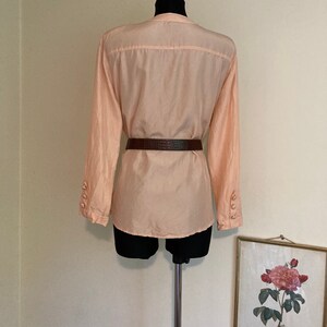 May include: A peach-colored long-sleeved blouse with a button-up front and a brown belt. The blouse is on a mannequin.
