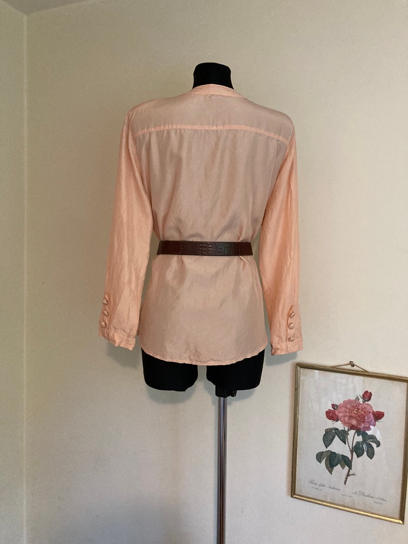 May include: A peach-colored long-sleeved blouse with a button-up front and a brown belt. The blouse is on a mannequin.