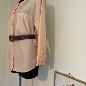 May include: A peach-colored silk blouse with a button-up front, long sleeves, and a single pocket on the left chest. The blouse is worn with a brown leather belt.
