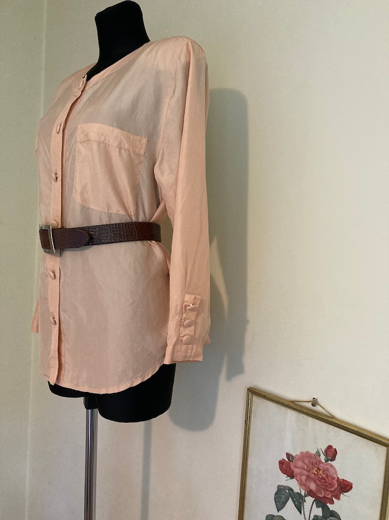 May include: A peach-colored silk blouse with a button-up front, long sleeves, and a single pocket on the left chest. The blouse is worn with a brown leather belt.
