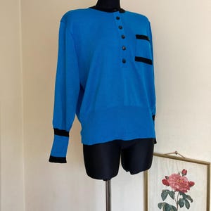 Peut inclure: Un pull bleu avec un col noir et des bordures noires sur les manches et la poche. Le pull a une patte de boutonnage et une seule poche sur le côté gauche.