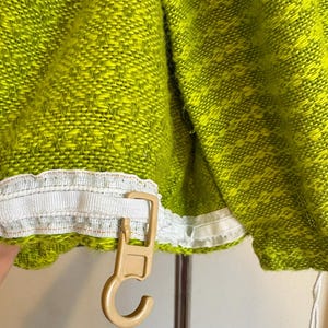 May include: Close-up of a lime green woven fabric with a white trim and a beige plastic hook. The fabric has a textured appearance with a pattern of small, interwoven threads. The hook is attached to the fabric's edge.