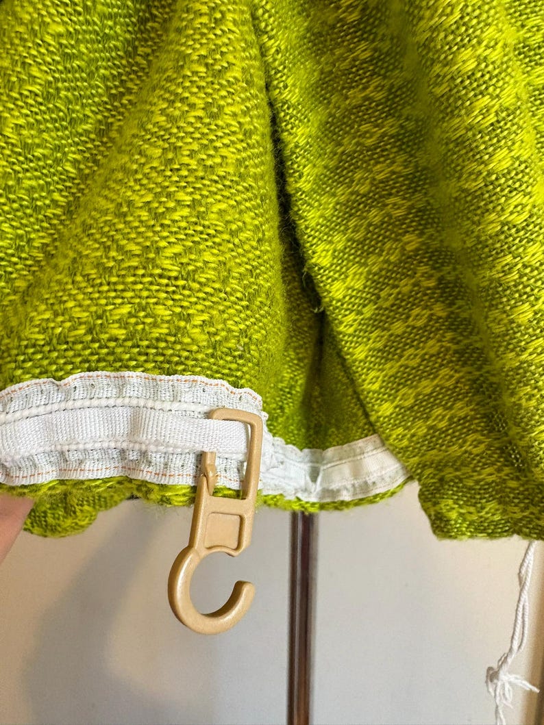 May include: Close-up of a lime green woven fabric with a white trim and a beige plastic hook. The fabric has a textured appearance with a pattern of small, interwoven threads. The hook is attached to the fabric's edge.