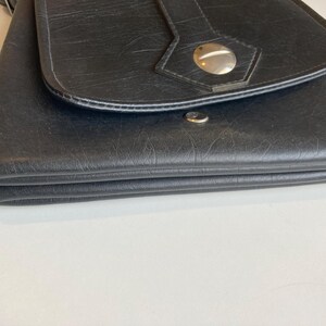 Pode incluir: Bolsa transversal de couro preto com fecho de aba. A bolsa apresenta uma superf&iacute;cie texturizada e um fecho de bot&atilde;o prateado. A al&ccedil;a &eacute; vis&iacute;vel na parte superior. A bolsa est&aacute; dobrada.