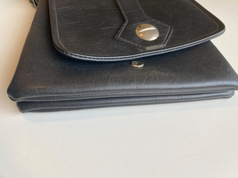 Pode incluir: Bolsa transversal de couro preto com fecho de aba. A bolsa apresenta uma superf&iacute;cie texturizada e um fecho de bot&atilde;o prateado. A al&ccedil;a &eacute; vis&iacute;vel na parte superior. A bolsa est&aacute; dobrada.