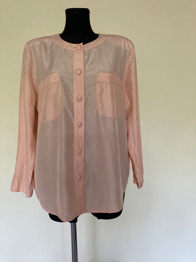 May include: A pale pink, long-sleeved button-up shirt with two patch pockets on the front. The shirt is made of a silky material.
