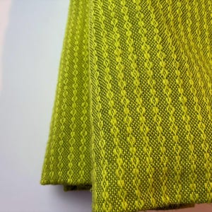 May include: Close-up of a folded, textured fabric in shades of green and yellow. The fabric has a diamond pattern. The material appears to be woven, with a soft, slightly fuzzy texture. The fabric is likely used for home decor.