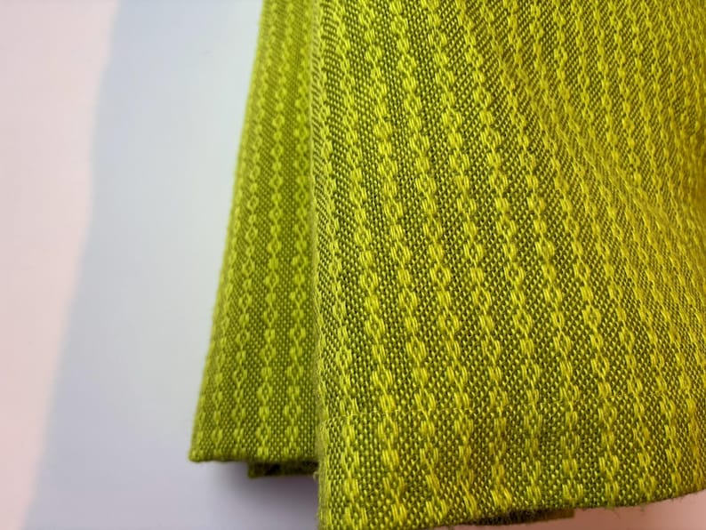 May include: Close-up of a folded, textured fabric in shades of green and yellow. The fabric has a diamond pattern. The material appears to be woven, with a soft, slightly fuzzy texture. The fabric is likely used for home decor.