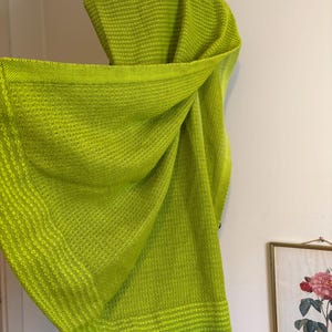 May include: A bright lime green woven shawl with a textured pattern. The shawl is draped, showcasing its soft, flowing fabric. The edges feature a contrasting, solid border. The shawl is displayed against a white wall.