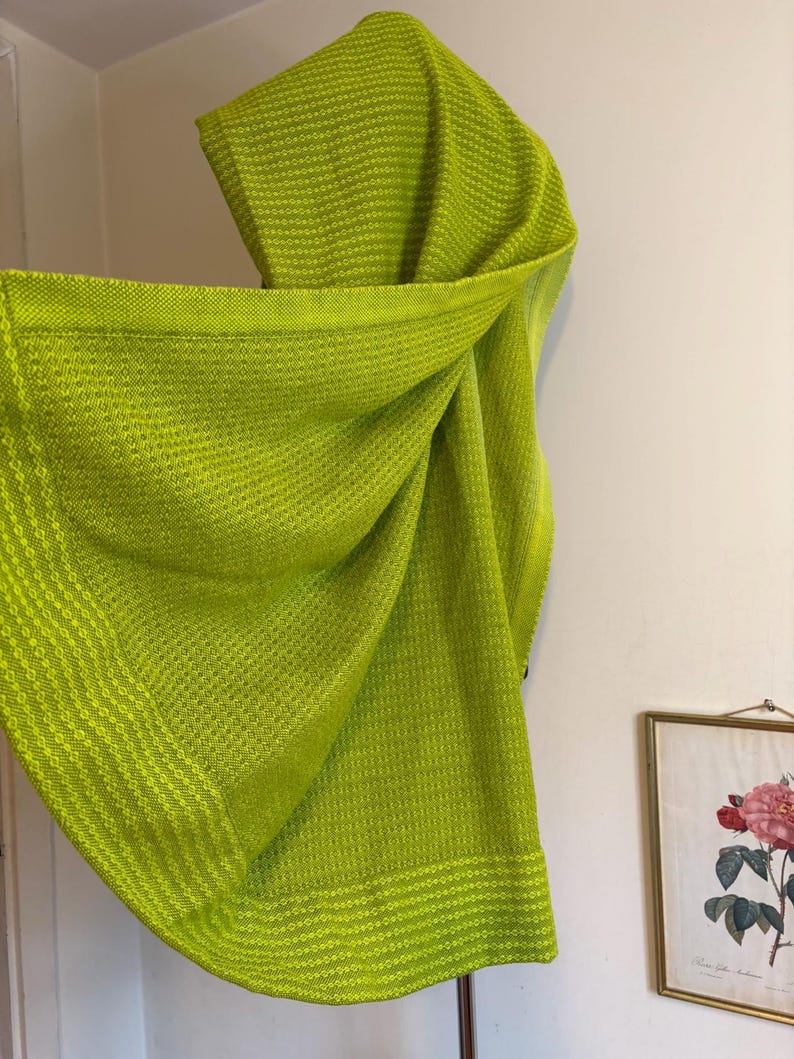May include: A bright lime green woven shawl with a textured pattern. The shawl is draped, showcasing its soft, flowing fabric. The edges feature a contrasting, solid border. The shawl is displayed against a white wall.
