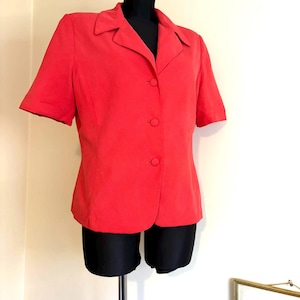 Puede incluir: Un blazer rojo brillante de manga corta con cuello clásico y tres botones a juego. El blazer se exhibe en un maniquí negro, mostrando su ajuste a medida y su color vibrante. La prenda está hecha de una tela suave.