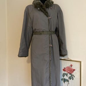 May include: A long gray coat with a faux fur collar and a belt. The coat has a zipper closure and a button detail. The coat is lined with a brown fabric.