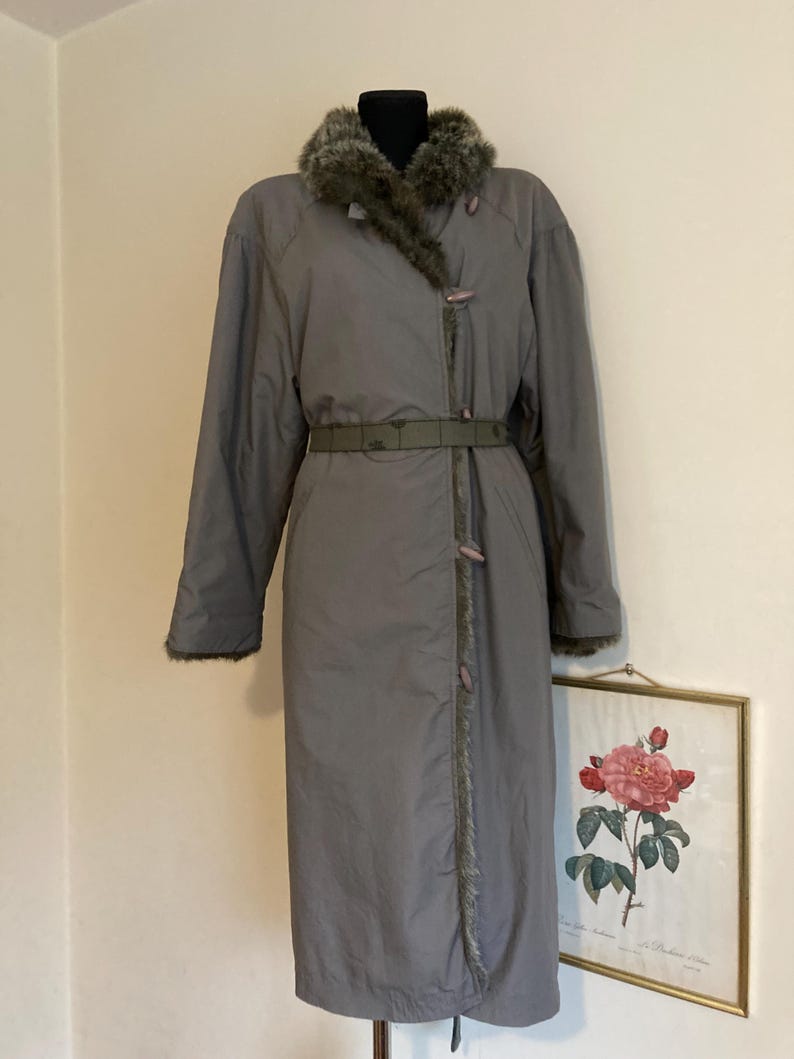 May include: A long gray coat with a faux fur collar and a belt. The coat has a zipper closure and a button detail. The coat is lined with a brown fabric.