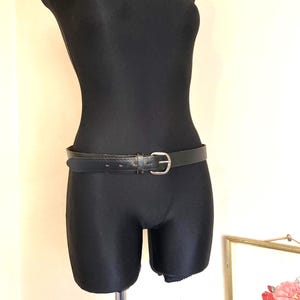 May include: A black leather belt with a silver buckle is fastened around the waist of a black mannequin. The belt has a simple, classic design, suitable for various outfits. The mannequin is on a metal stand.