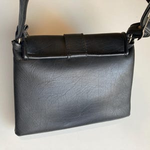 Pode incluir: Uma bolsa transversal preta com uma superf&iacute;cie texturizada. A bolsa possui um fecho de aba com fivela e uma al&ccedil;a de ombro ajust&aacute;vel. A bolsa &eacute; feita de material sint&eacute;tico.