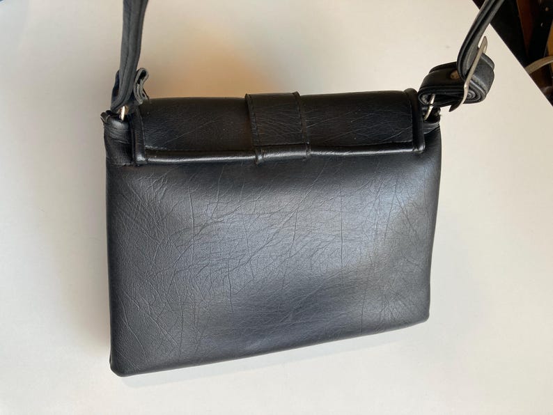 Pode incluir: Uma bolsa transversal preta com uma superf&iacute;cie texturizada. A bolsa possui um fecho de aba com fivela e uma al&ccedil;a de ombro ajust&aacute;vel. A bolsa &eacute; feita de material sint&eacute;tico.