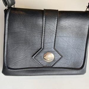Pode incluir: Bolsa transversal preta com acabamento texturizado. A bolsa possui fecho de aba com bot&atilde;o prateado e al&ccedil;a decorativa. Possui al&ccedil;a de ombro e &eacute; feita de material semelhante ao couro.