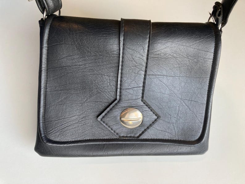 Pode incluir: Bolsa transversal preta com acabamento texturizado. A bolsa possui fecho de aba com bot&atilde;o prateado e al&ccedil;a decorativa. Possui al&ccedil;a de ombro e &eacute; feita de material semelhante ao couro.
