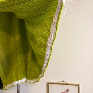 May include: Close-up of a lime green woven fabric with a white ribbon trim. The fabric has a textured pattern and is hanging from a support. The edge of the fabric is visible, showing the trim detail.
