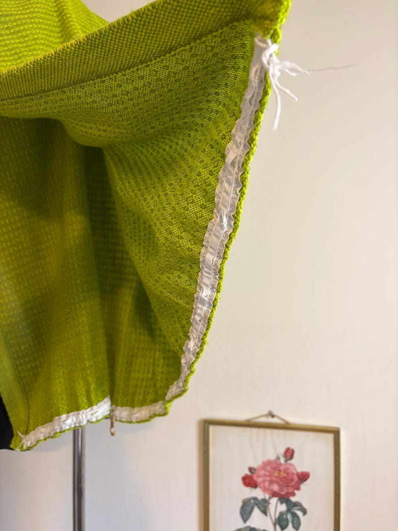 May include: Close-up of a lime green woven fabric with a white ribbon trim. The fabric has a textured pattern and is hanging from a support. The edge of the fabric is visible, showing the trim detail.