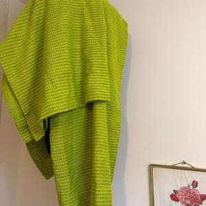 May include: A lime green woven poncho with a textured pattern hangs against a white wall. The poncho is draped over a hanger, showcasing its shape and design. A framed floral print is visible in the background.