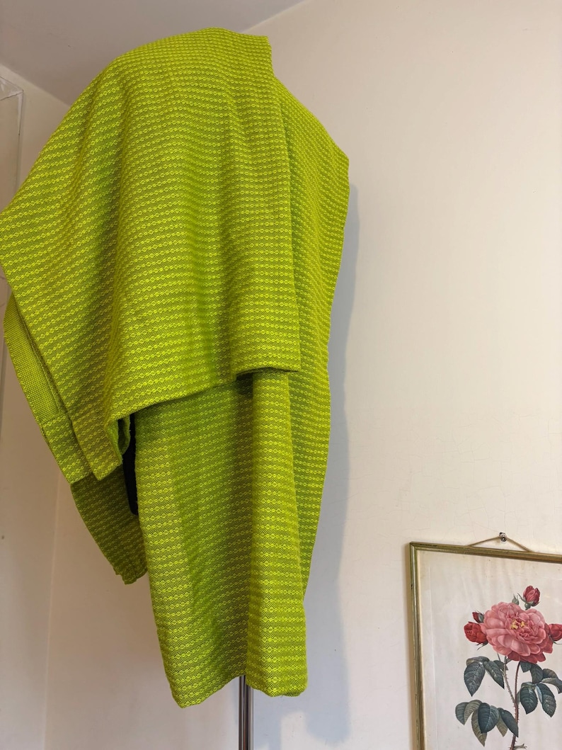 May include: A lime green woven poncho with a textured pattern hangs against a white wall. The poncho is draped over a hanger, showcasing its shape and design. A framed floral print is visible in the background.