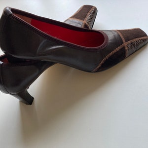 May include: A pair of brown leather heeled shoes with a suede and patterned design. The shoes have a pointed toe and a low heel.
